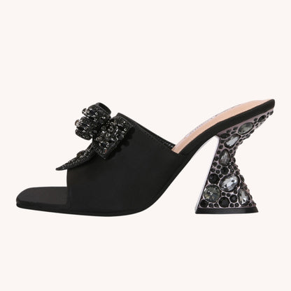 A black satin mule sandal with a chunky block heel embellished with dark gemstones and a matching beaded bow detail on the strap, shown against a solid off-white background.