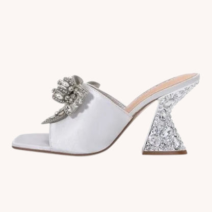 A single white satin mule sandal with a chunky high heel covered in clear rhinestones, featuring a matching rhinestone-embellished bow on the strap, shown against a solid off-white background.