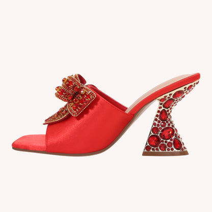 A red satin mule sandal with a decorative red and gold crystal bow on the strap and a sculptural high heel covered in red crystals, shown against a light cream background.