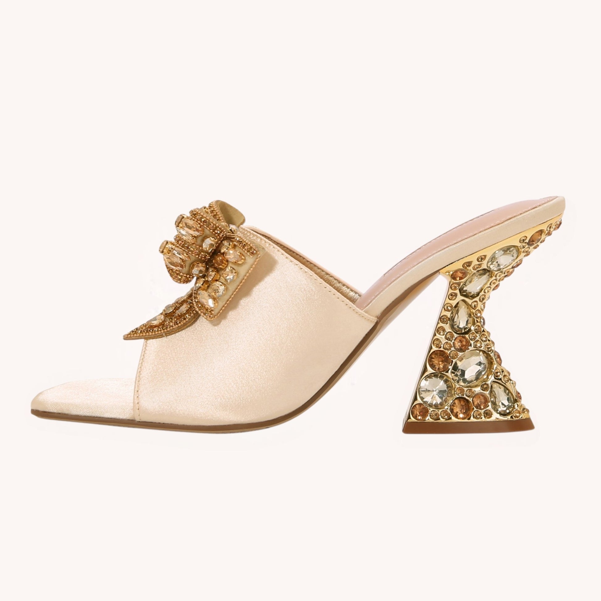 A single pale blush satin mule sandal with a sculptural gold and crystal embellished block heel and a matching jeweled bow detail on the strap, shown against a solid off-white background.